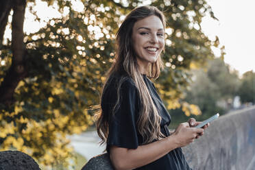 Happy young woman with long hair holding smart phone by wall - JOSEF13479