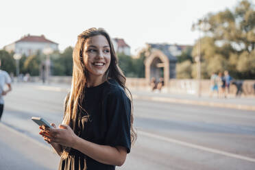 Lächelnde junge Frau mit Mobiltelefon auf der Straße stehend - JOSEF13480