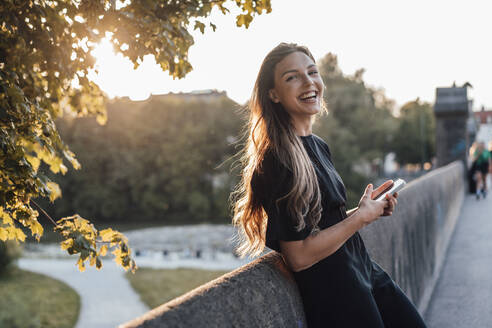 Cheerful woman with smart phone leaning on wall - JOSEF13481