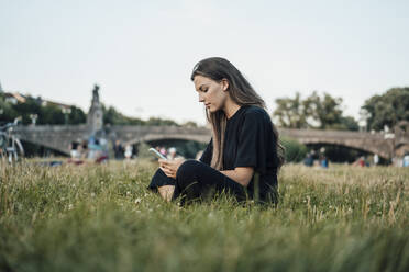 Woman text messaging through mobile phone sitting in park - JOSEF13483
