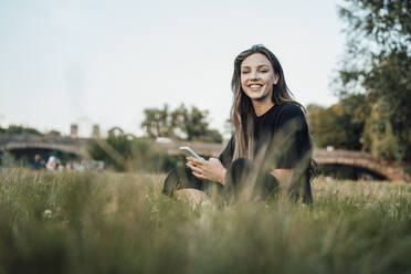 Happy woman with mobile phone sitting in park - JOSEF13484