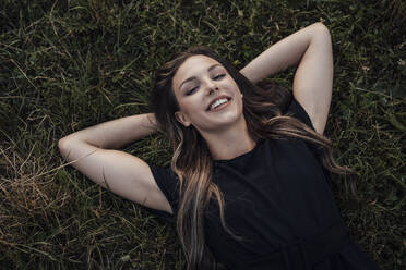 Happy young woman with hands behind head relaxing on grass at park - JOSEF13486