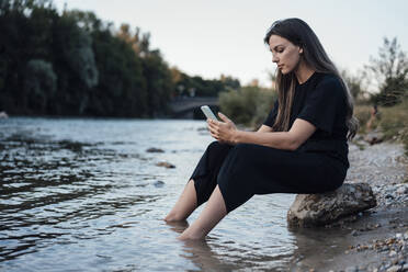 Junge Frau mit Mobiltelefon auf einem Felsen am See sitzend - JOSEF13491