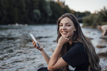 Glückliche junge Frau mit Handy in der Hand am See sitzend mit Hand am Kinn - JOSEF13493