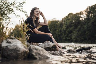 Lächelnde, nachdenkliche junge Frau, die am See sitzt - JOSEF13499