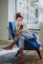 Lächelnde Frau mit Smartphone auf einem Stuhl zu Hause - JOSEF13533