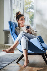 Smiling thoughtful woman with smart phone on chair at home - JOSEF13537