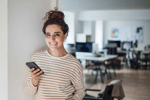 Lächelnde Geschäftsfrau mit Smartphone an der Wand im Büro - JOSEF13551