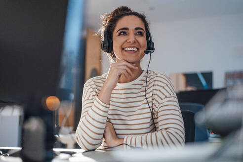 Happy businesswoman with hand on chin using headset in office - JOSEF13555