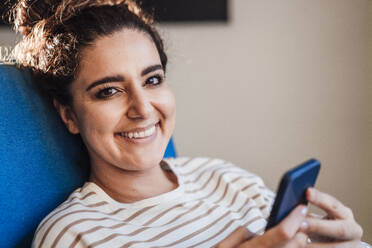 Smiling woman with smart phone on chair at home - JOSEF13577