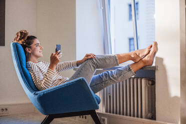 Nachdenkliche Frau mit Handy entspannt sich auf einem Stuhl zu Hause - JOSEF13580