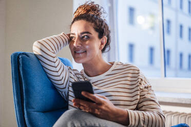 Smiling woman with mobile phone contemplating on chair at home - JOSEF13581