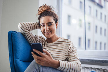 Smiling woman with mobile phone on chair at home - JOSEF13582