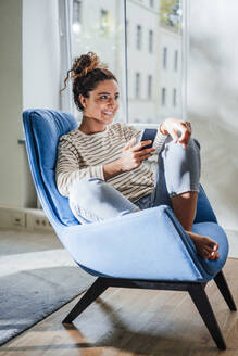 Lächelnde Frau mit Mobiltelefon auf einem Stuhl sitzend zu Hause - JOSEF13584