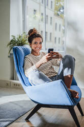 Smiling woman with mobile phone on blue chair at home - JOSEF13586