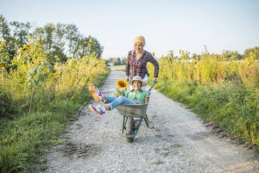 Happy gardener pushing colleague sitting in wheelbarrow at field - AANF00335