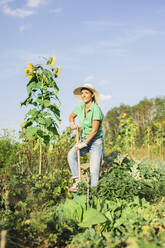 Lächelnder reifer Gärtner mit Schaufel bei der Gartenarbeit im Feld an einem sonnigen Tag - AANF00340