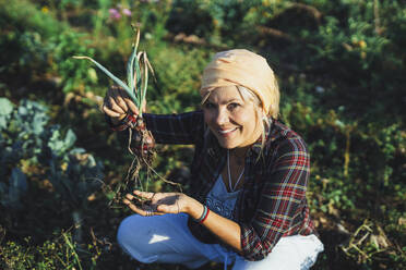 Happy mature gardener holding scallion crouching at field - AANF00349