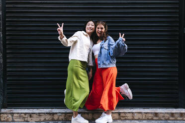 Cheerful lesbian couple showing peace sign in front of shutter - ASGF02915