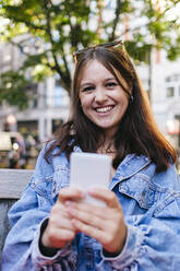 Happy woman wearing denim jacket holding mobile phone - ASGF02932