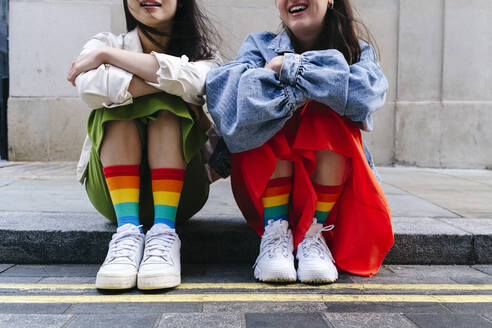 Lesbian women wearing shoes and multi colored socks sitting on footpath - ASGF02966