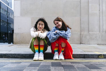 Smiling lesbian friends with multi colored socks sitting on footpath - ASGF02968