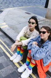 Young gay couple wearing sunglasses sitting on footpath - ASGF02970