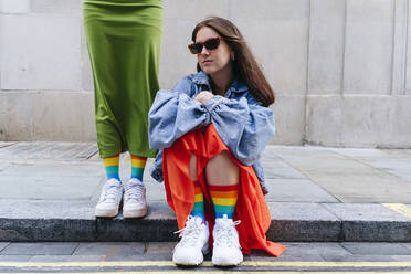 Woman wearing sunglasses sitting by lesbian friend standing on footpath - ASGF02971