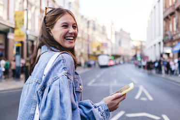 Cheerful young woman holding mobile phone strolling in city - ASGF02988