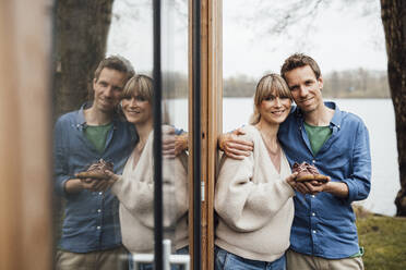 Smiling expectant parents holding baby booties by glass reflection - JOSEF13609