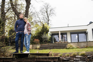 Expectant couple standing on jetty in front of house - JOSEF13618