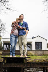 Happy expectant couple embracing each other standing on jetty in front of house - JOSEF13630