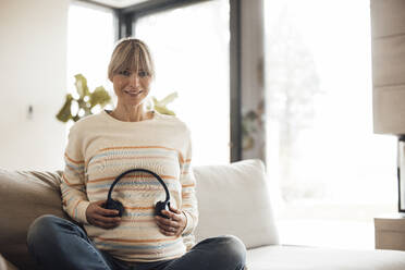 Smiling woman holding wireless headphones on pregnant belly at home - JOSEF13635