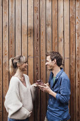 Pregnant woman holding baby booties with man in front of wooden wall - JOSEF13680