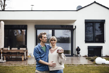 Man touching belly of pregnant woman holding baby booties in front of house - JOSEF13681