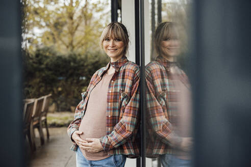 Smiling pregnant woman touching belly leaning on glass door - JOSEF13688