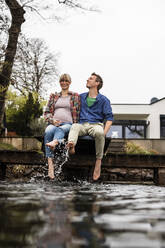 Verspieltes schwangeres Paar, das auf einem Steg am See sitzt und Wasser spritzt - JOSEF13709