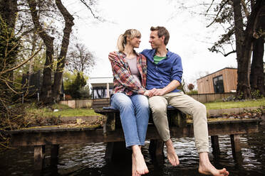 Romantic expectant couple looking at each other sitting on jetty - JOSEF13710