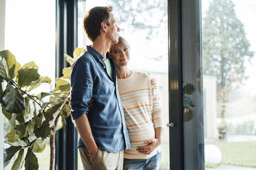 Mature man standing with pregnant woman near glass door at home - JOSEF13716