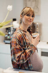 Nachdenkliche schwangere Frau in kariertem Hemd mit Tasse zu Hause - JOSEF13726