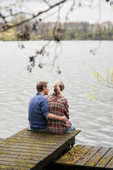 Man with arm around woman sitting on jetty at lake - JOSEF13734