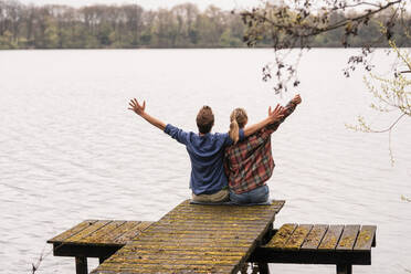 Carefree couple with arms outstretched sitting on jetty at lake - JOSEF13736