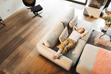 Relaxed woman with hands behind head lying on sofa in living room at home - JOSEF13743