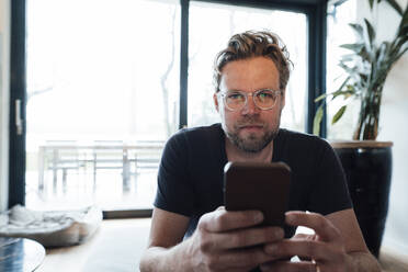 Mature man wearing eyeglasses holding smart phone at home - JOSEF13767