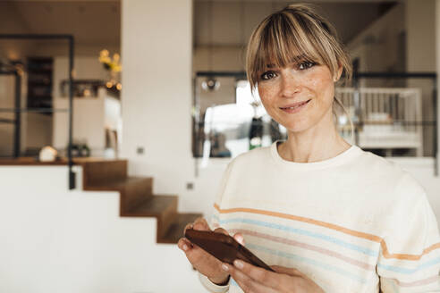 Smiling woman with bangs holding mobile phone at home - JOSEF13773