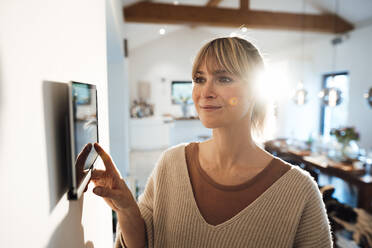 Smiling woman using smart home app through tablet PC mounted on wall at home - JOSEF13776