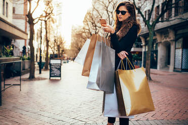 Shopper Frau Einkaufen mit einem Smartphone in einer Stadt Straße. Kaukasische Frau steht entlang der Straße mit Einkaufstaschen und mit Handy. - JLPSF01700