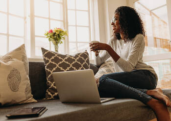 Businesswoman working on laptop computer sitting at home holding a coffee cup in hand. Smiling woman sitting on sofa at home and looking out of the window while working on laptop. - JLPSF01716