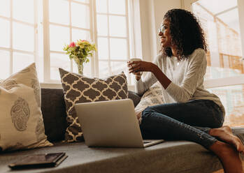 Geschäftsfrau, die an einem Laptop arbeitet, sitzt zu Hause und hält eine Kaffeetasse in der Hand. Lächelnde Frau, die zu Hause auf dem Sofa sitzt und aus dem Fenster schaut, während sie am Laptop arbeitet. - JLPSF01716