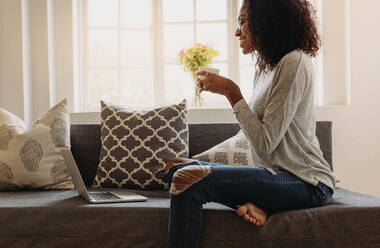 Lächelnde Frau in modischen zerrissenen Jeans hält eine Kaffeetasse, während sie zu Hause am Laptop arbeitet. Geschäftsfrau sitzt zu Hause auf dem Sofa und arbeitet am Laptop. - JLPSF01718